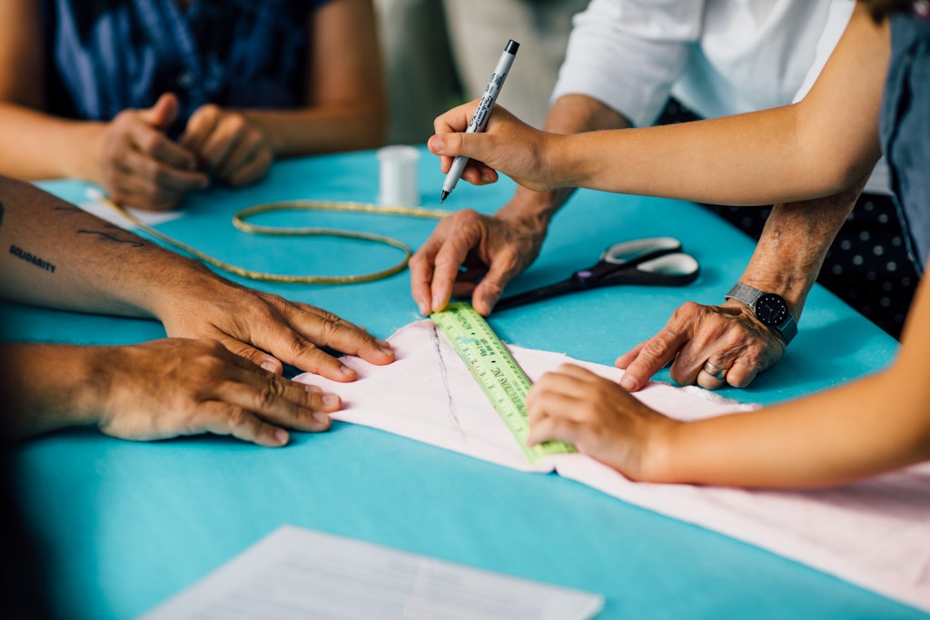 People measuring and marking fabric with a ruler and pen on a blue table, preparing for sewing.