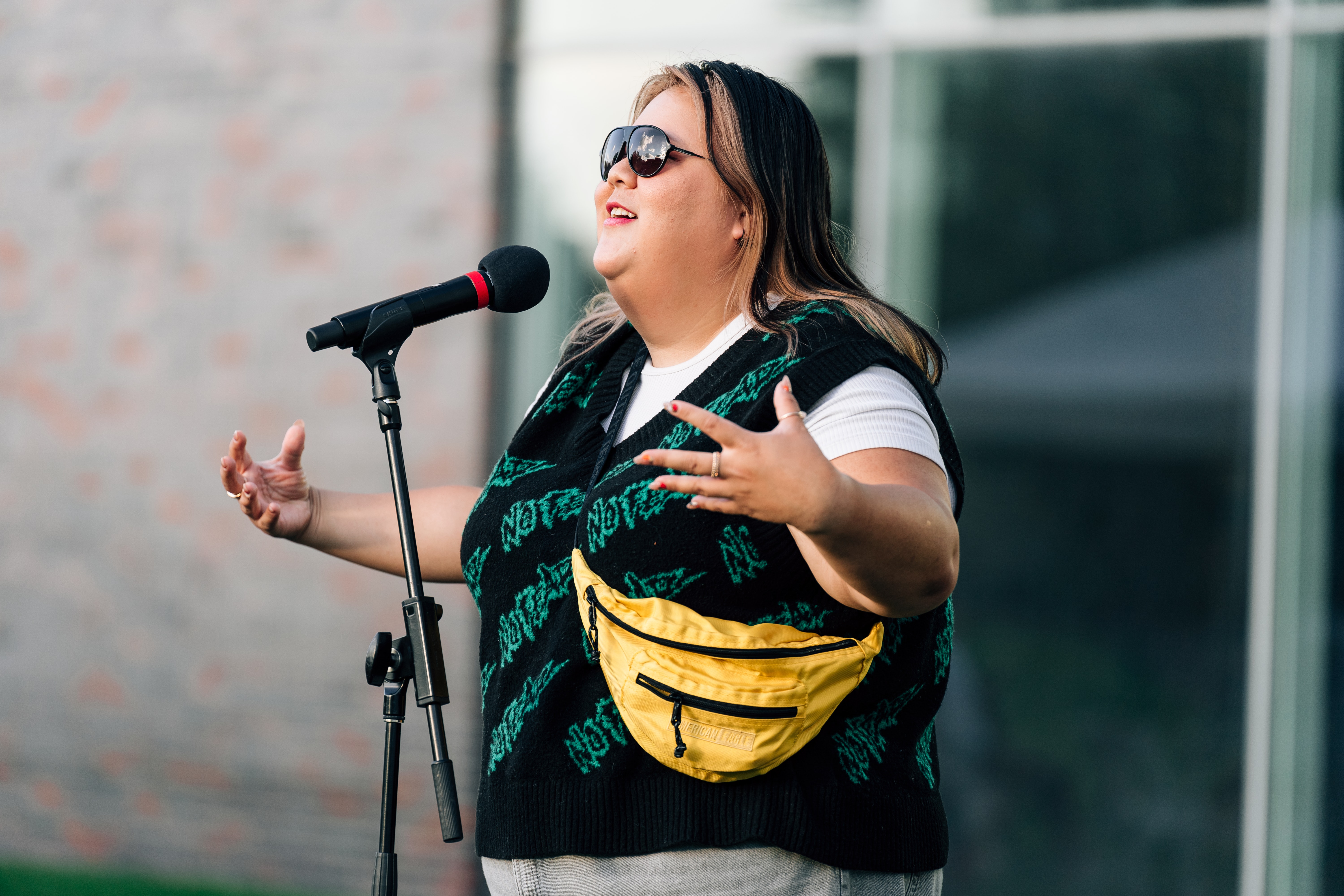 A woman wearing sunglasses and a yellow fanny pack speaks passionately into a microphone outdoors.