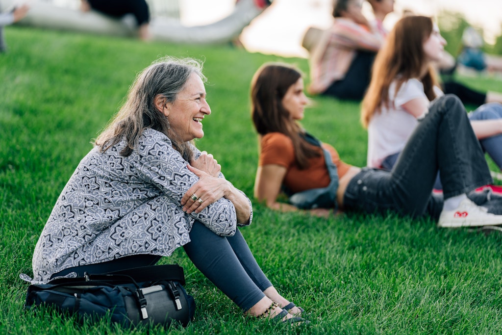 A light skinned adult with long grey hair sits in a grassy park among a crowd and smiles looking out.