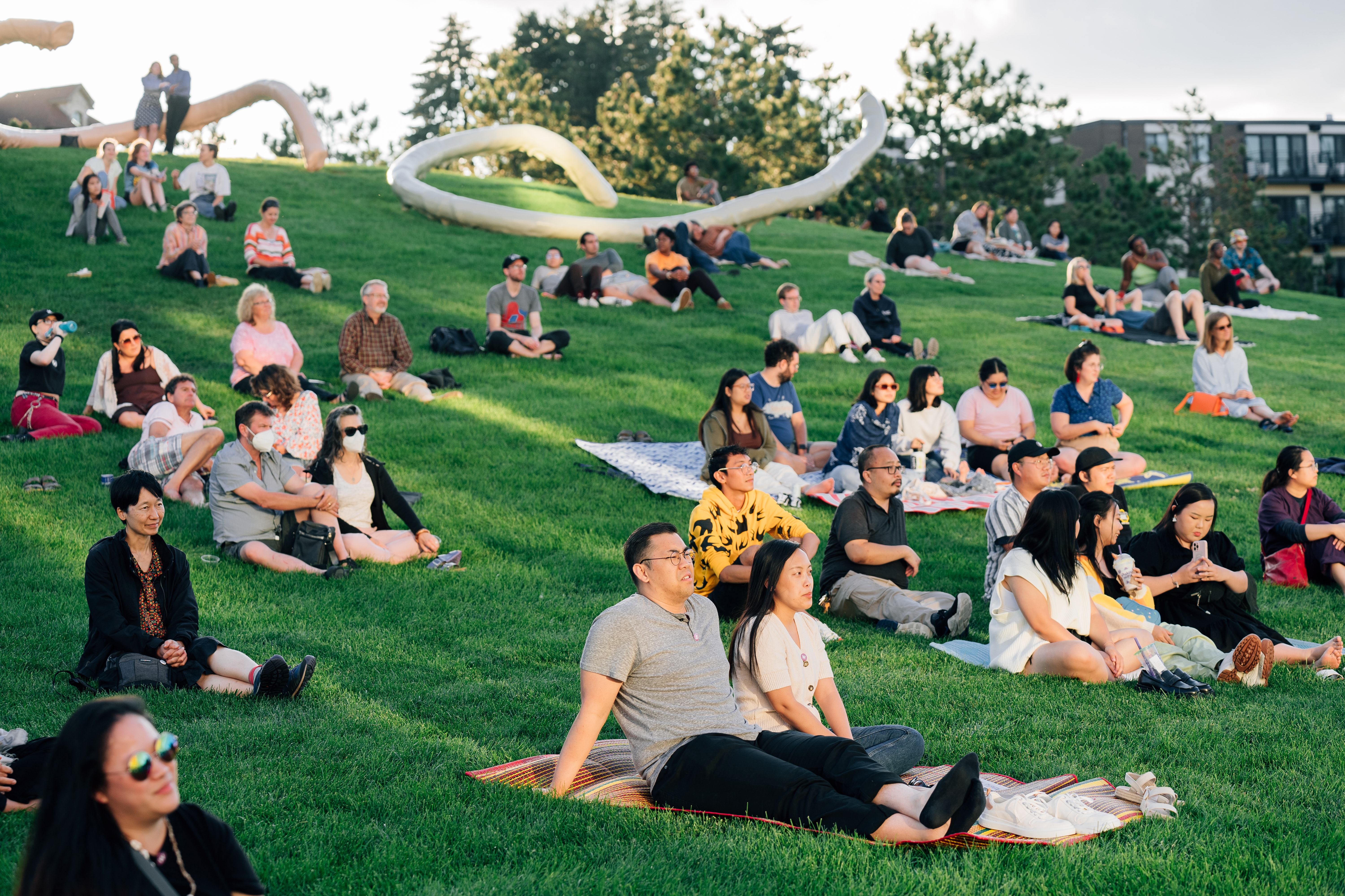 Adults sit on a hillside outdoors and listening to someone off camera.
