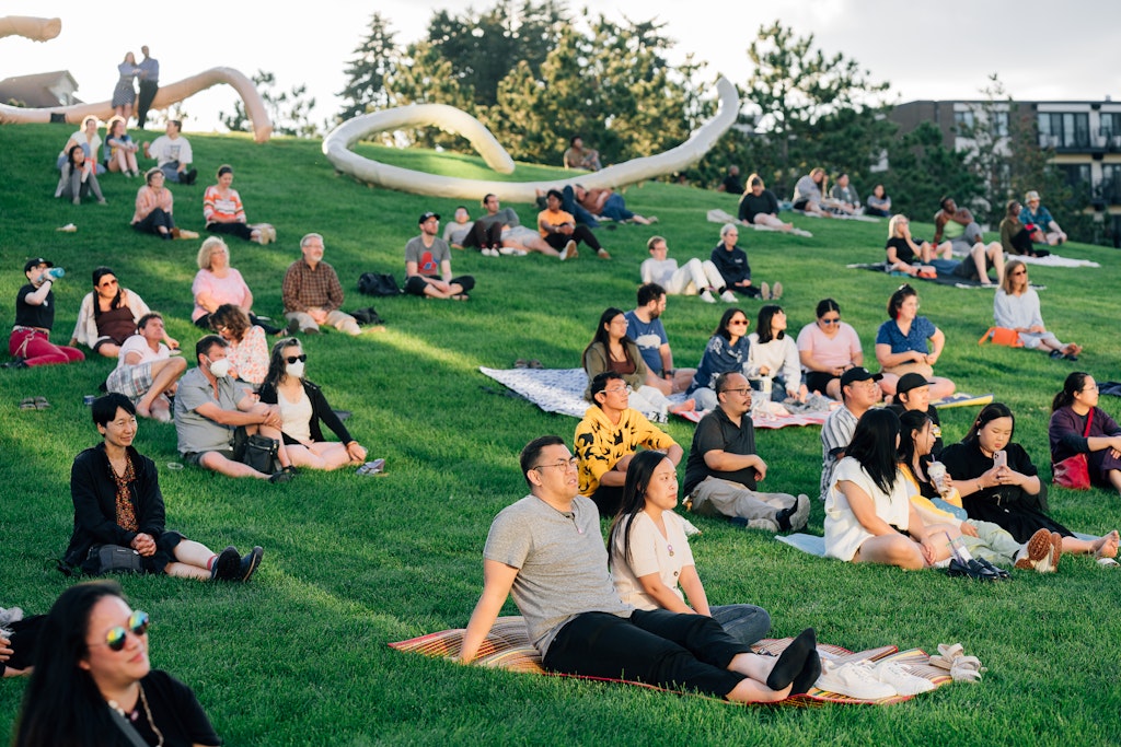 Adults sit on a hillside outdoors and listening to someone off camera.
