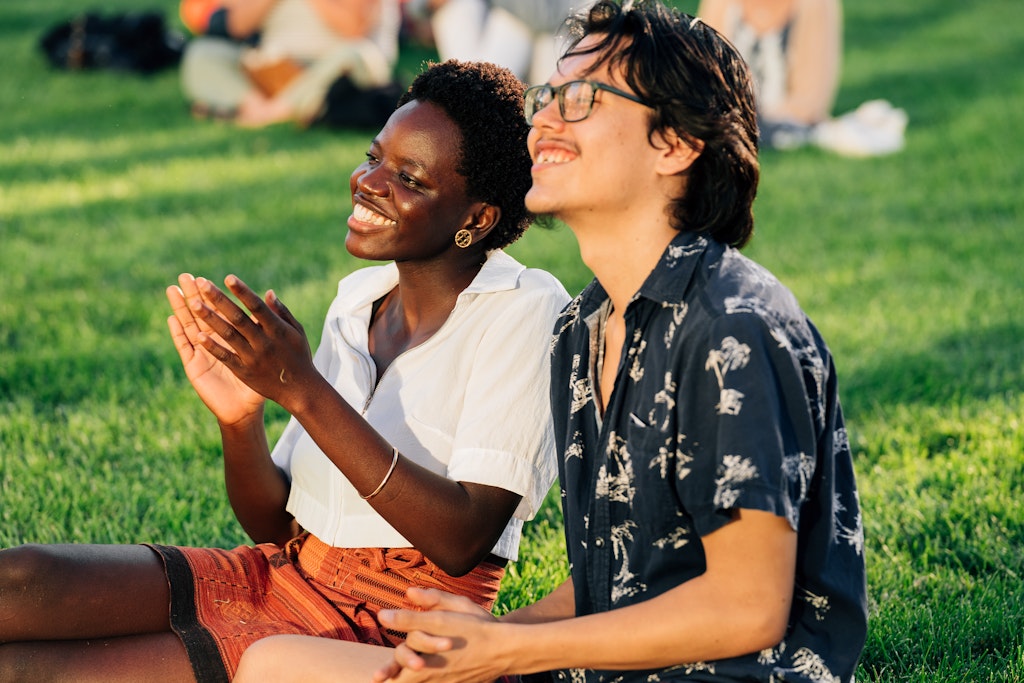 Two adults sit in a grassy park, smile and clap facing to the left.