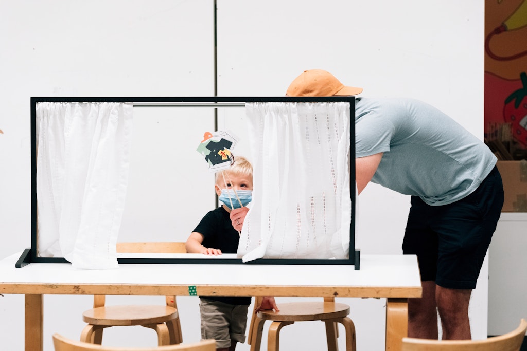 A child with a mask does a puppet show while an adult assists, both behind a small curtained stage.