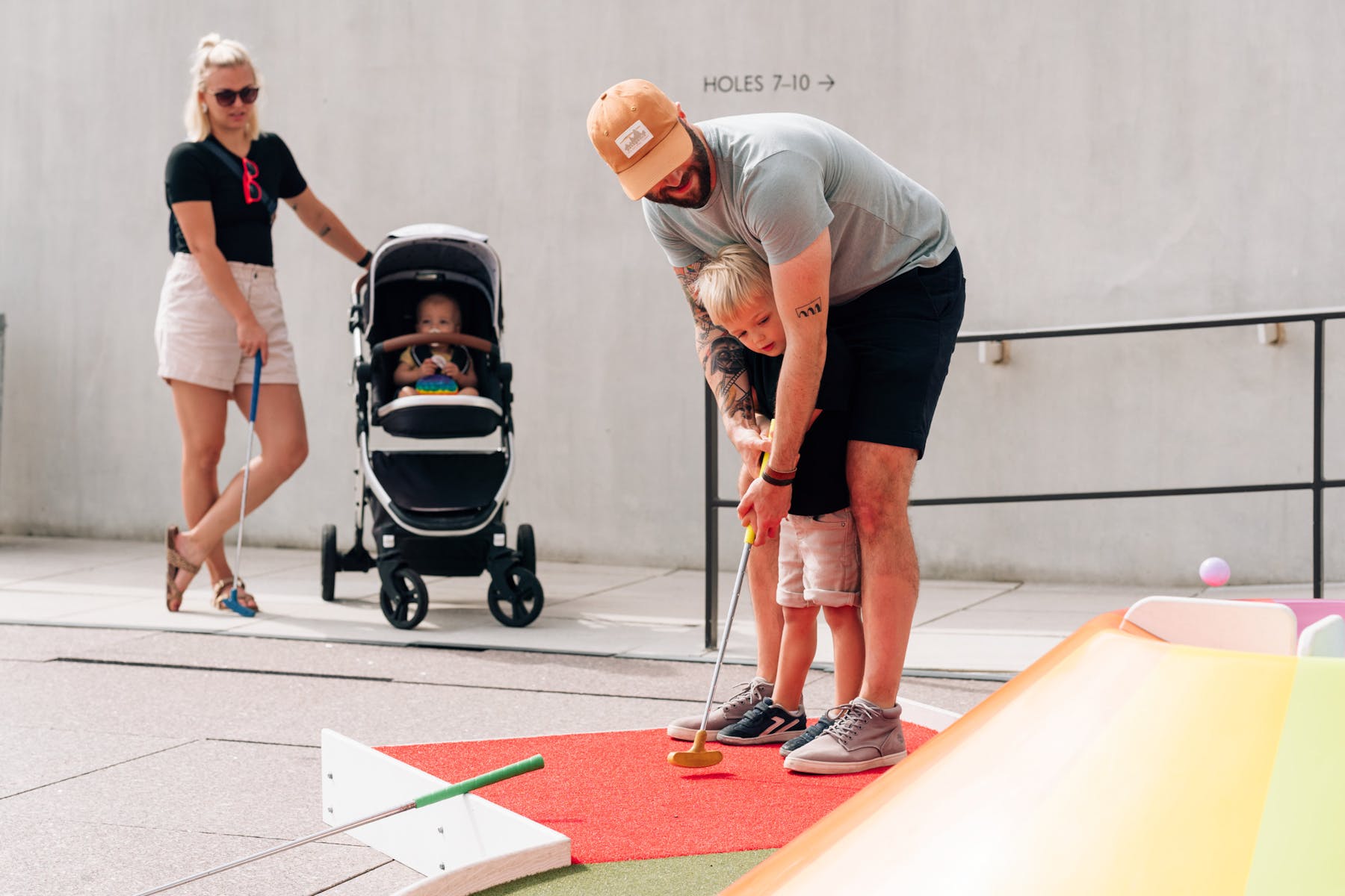 An adult playing mini golf with a child. An adult and a baby in a stroller in the background.