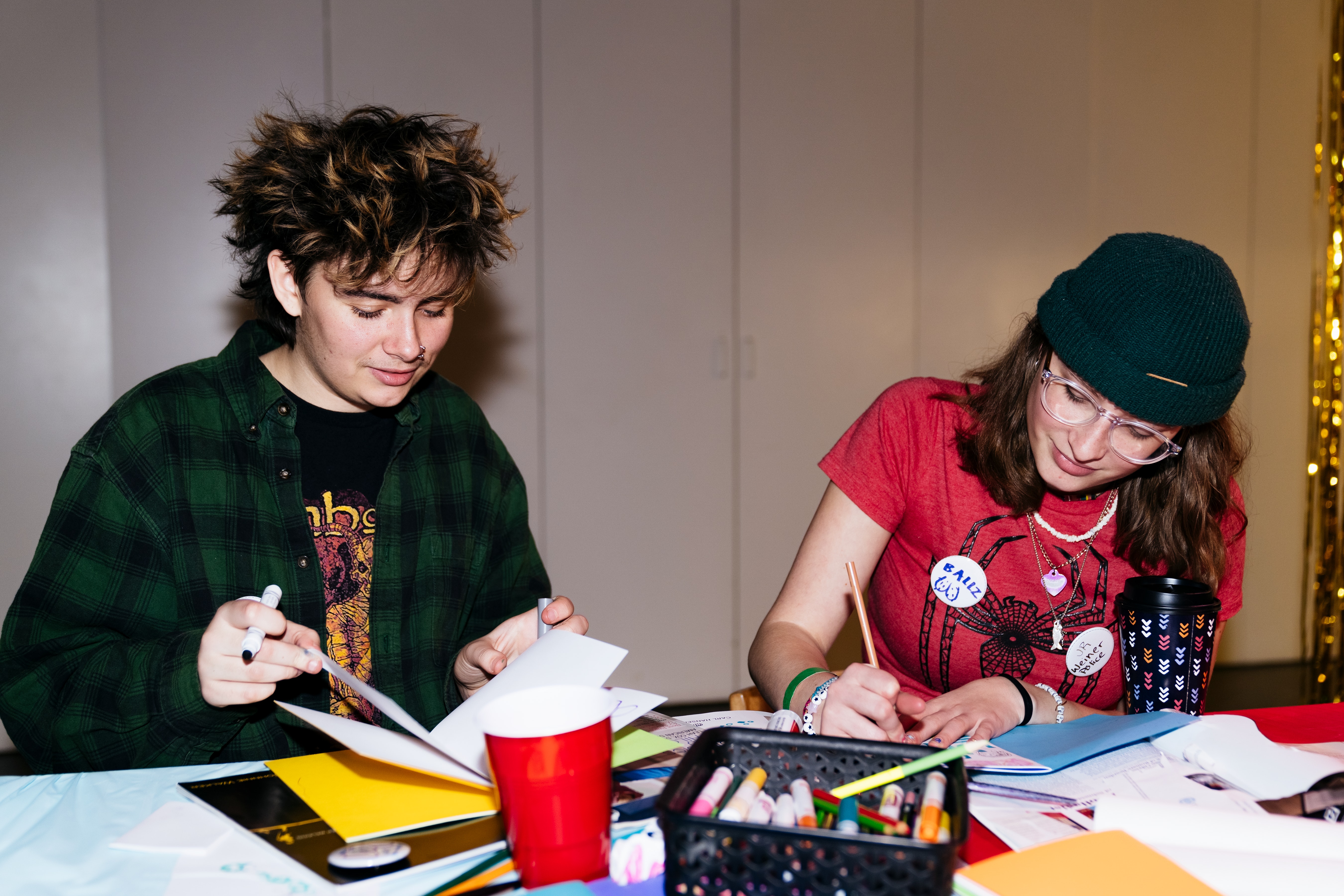 Two teenagers make notebook sized artwork at a table.