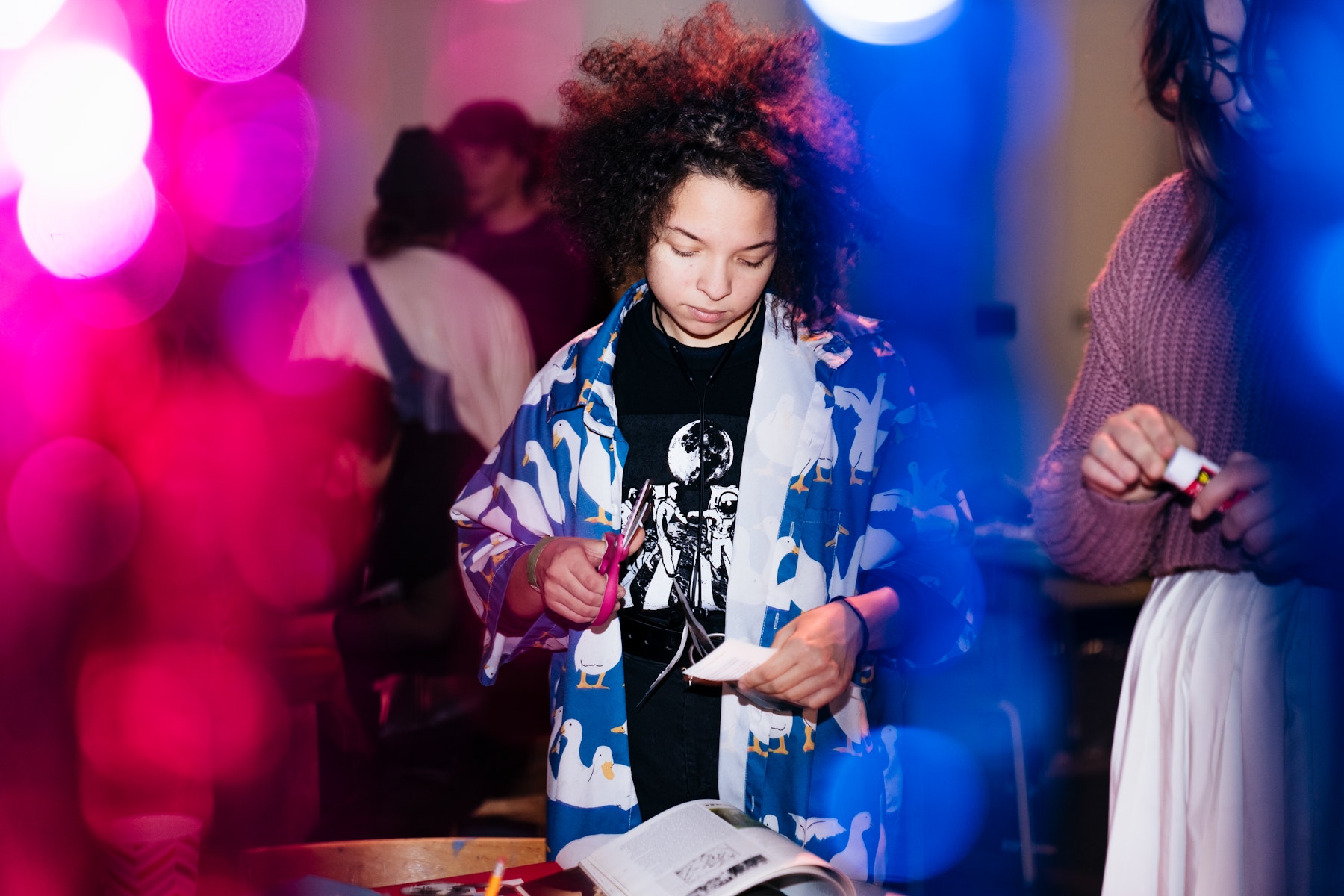 A teen stands in front of colorful streamers while making an artwork from paper and scissors at a table.
