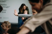People in a room, one is behind a blurring person i the forground and holds up an arm as if they are dancing.