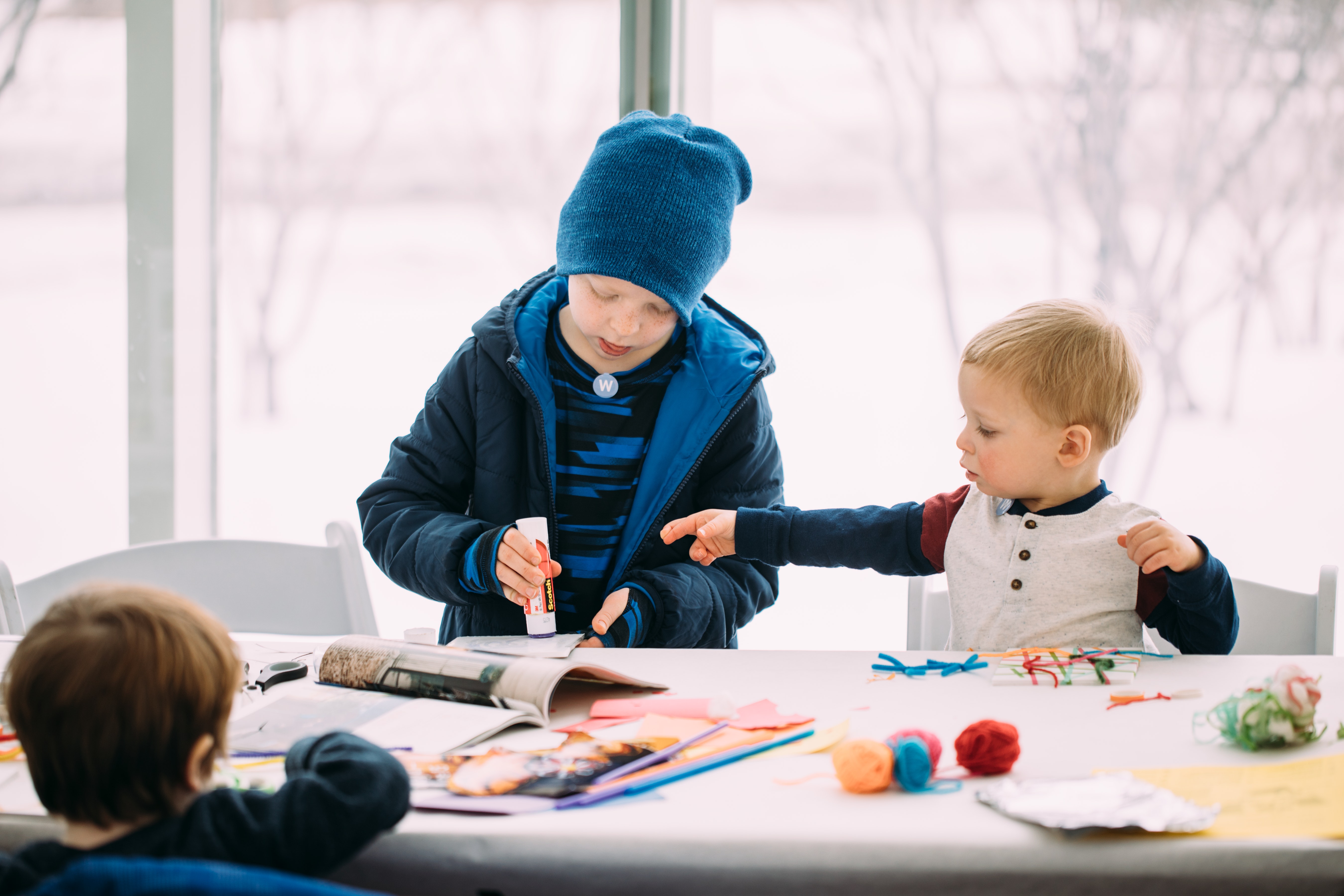 Two children with light skin make art at a table in front of large windows that look out over a snow covered garden.