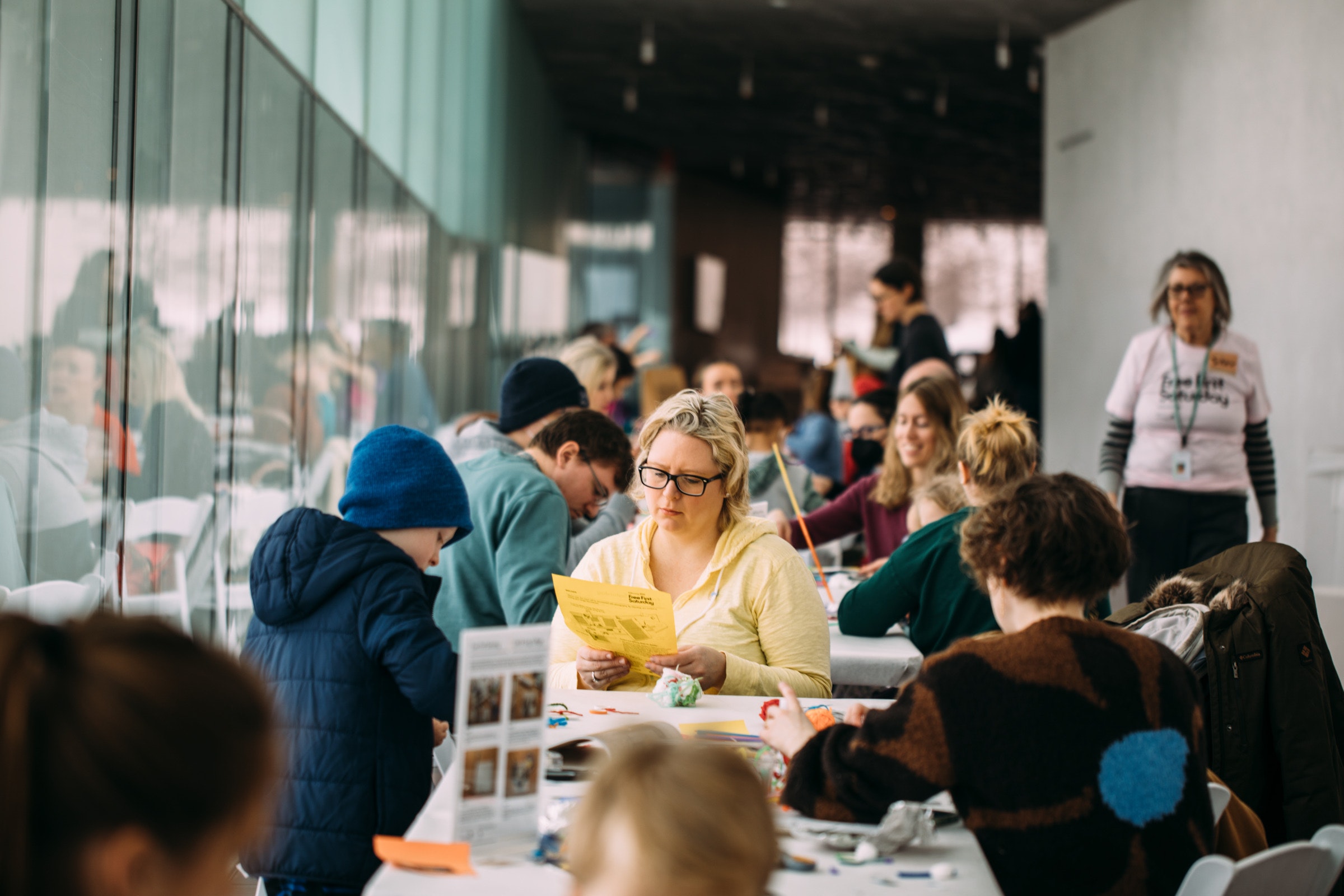 A crowd of people sit at tables working on crafts in a large space with windows to the left of frame.