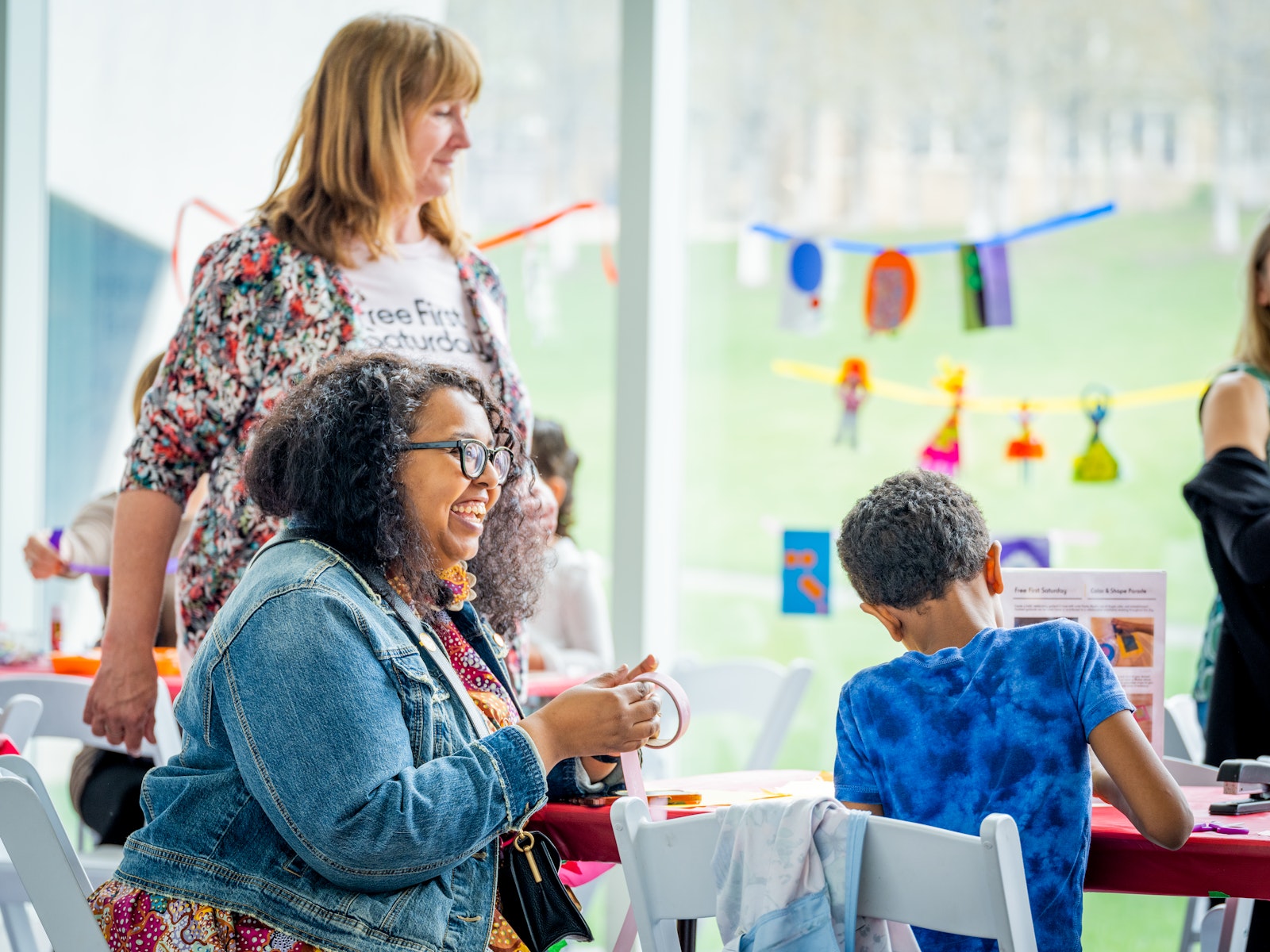 An adult with dark skin laughs and smiles as they sit at a table making artowrk with a child with dark skin while an adult with light skin stands nearby wearing a t-short that says "Free First Saturdays."