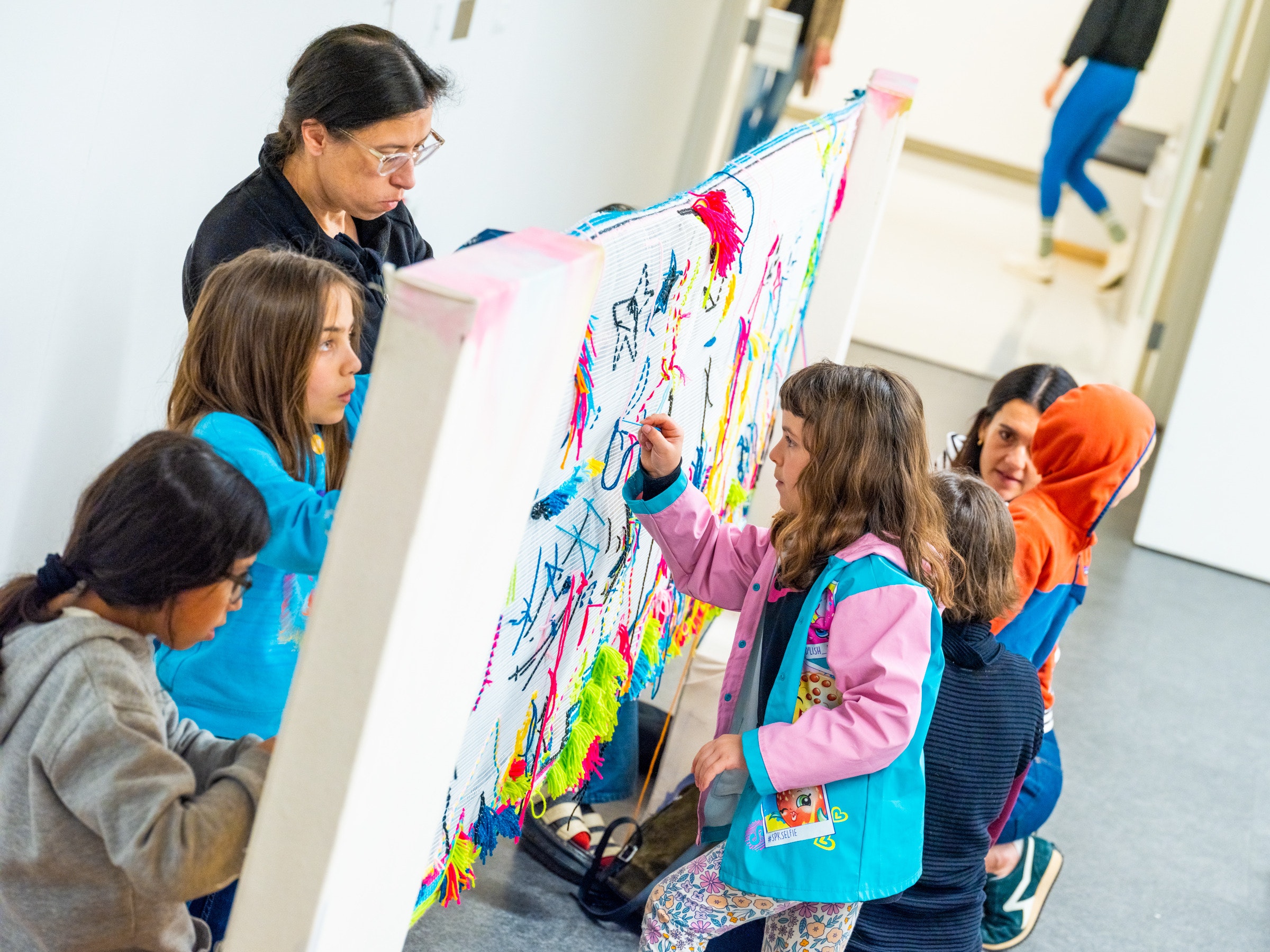 A group of children and adults work on a large piece of fabric stretched between two poles in a classroom.