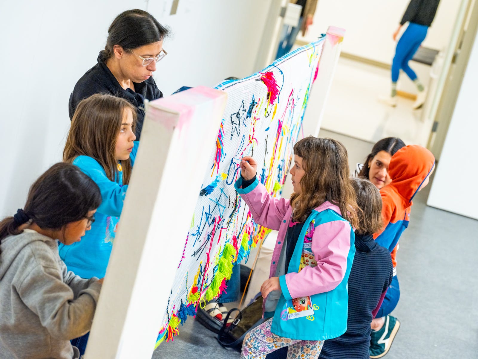 A group of children and adults work on a large piece of fabric stretched between two poles in a classroom.
