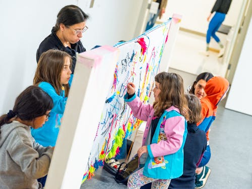 A group of children and adults work on a large piece of fabric stretched between two poles in a classroom.