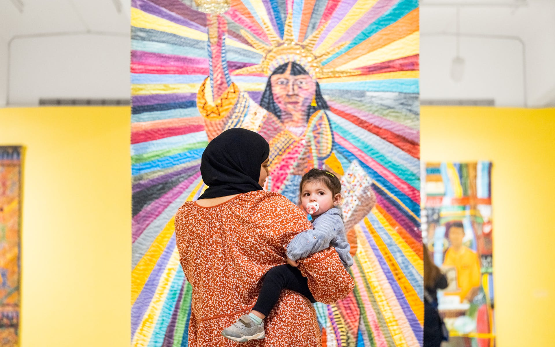 With their back to the viewer, a woman wearing a head covering holds a baby in her arms as she looks at a painting of the stature of liberty depicted as brown skined while the baby looks over its shoulder at the viewer.