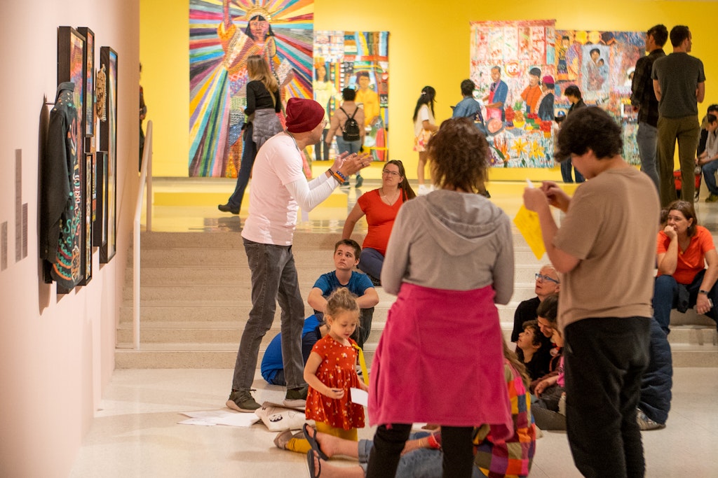An adult speaks to a group of adults and children in a colorful art gallery.