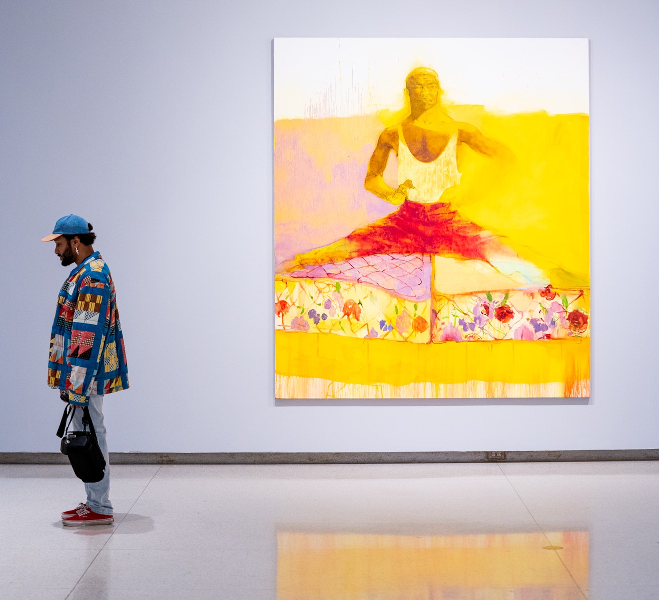 An adult with dark skin stands in an art gallery next to a large painting of an adult with dark skin in dancer clothing.