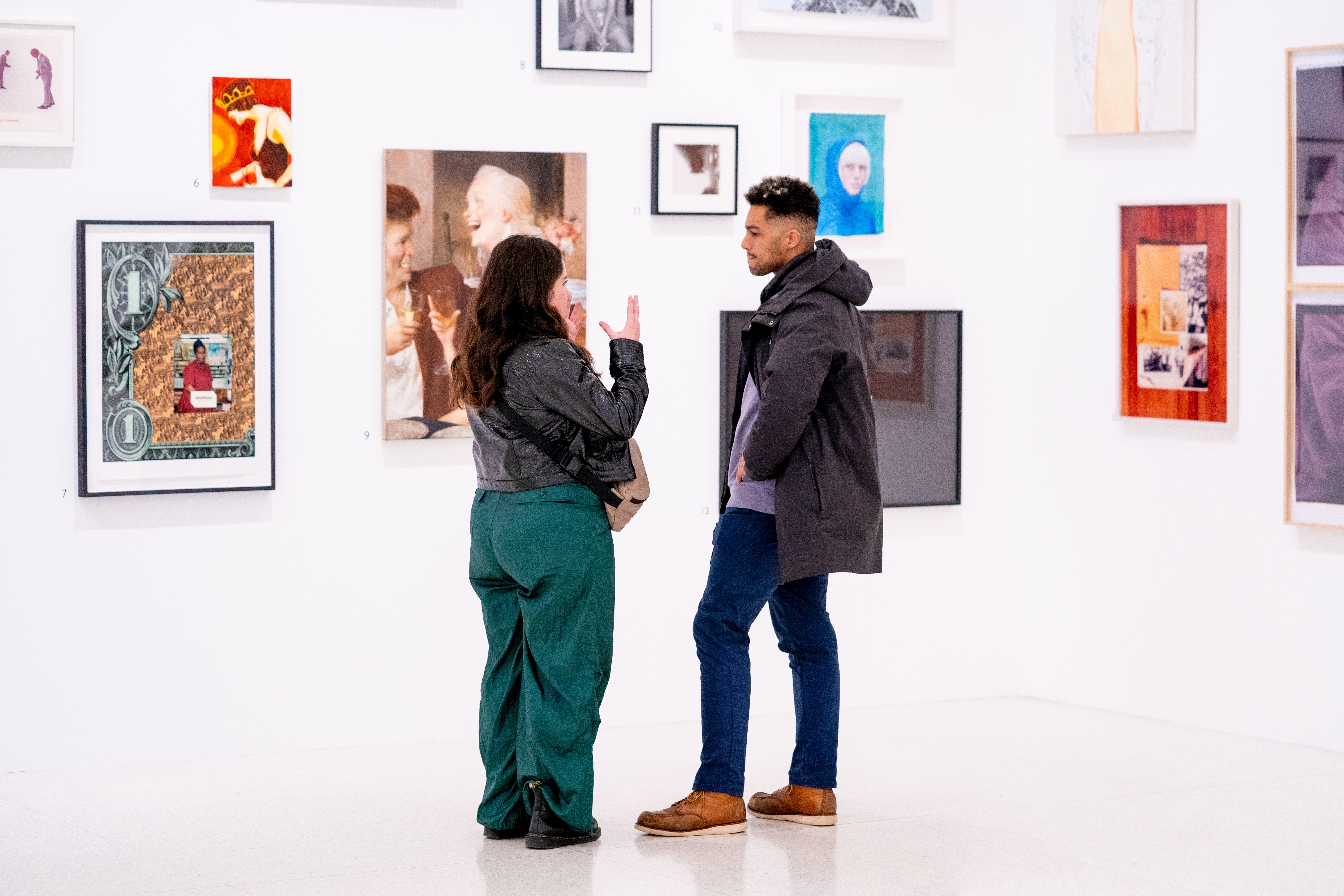Two adults talk to one another inf ront of a wall filled with portraits in a large white art gallery.