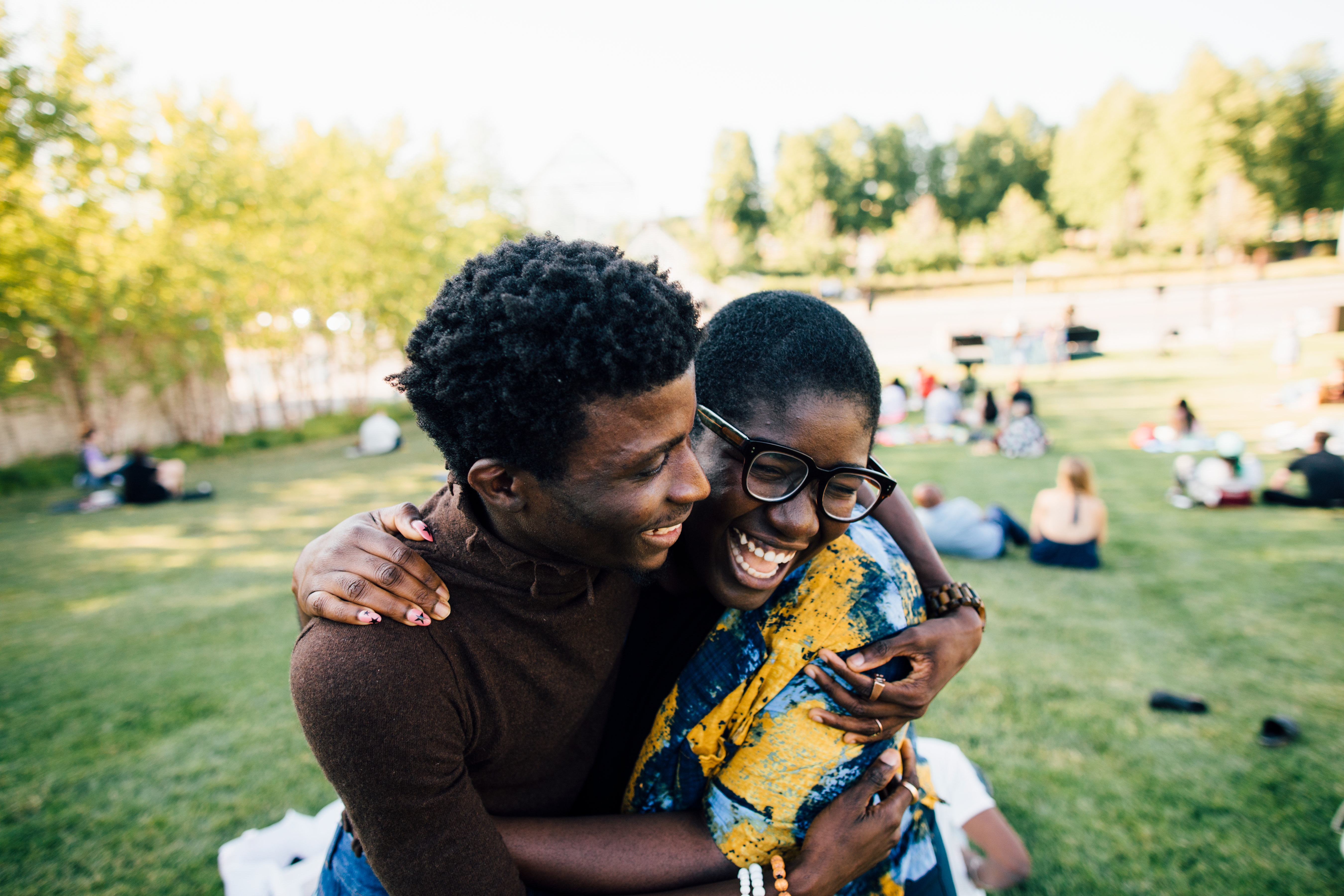 Two dark skinned adults embrace, smiling while sitting in a grassy park