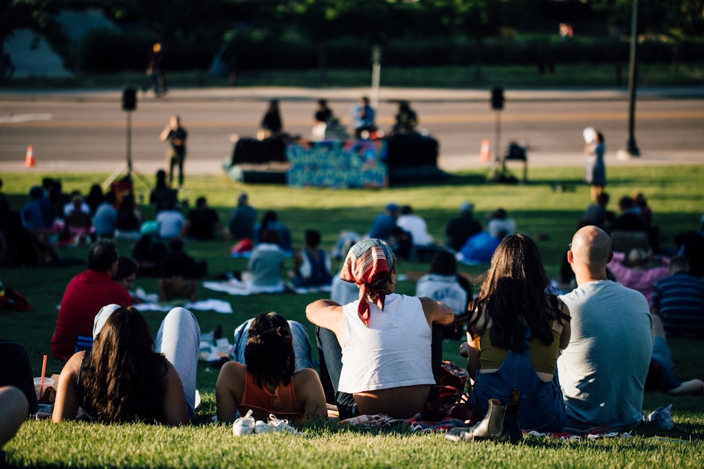 People sitting on grass at an outdoor concert, watching musicians perform on a stage in the distance.