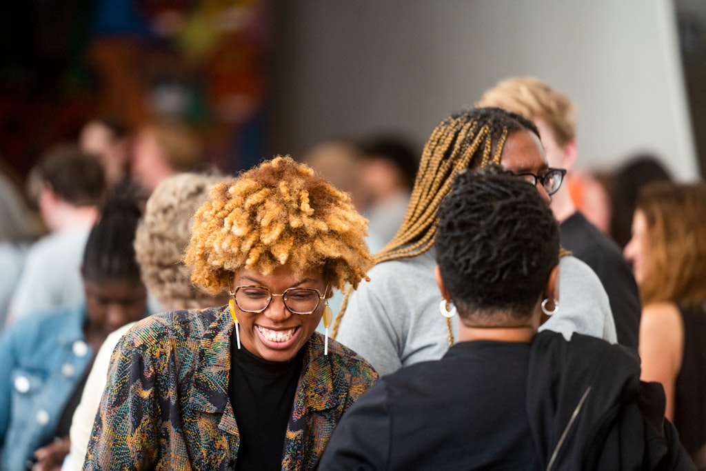A woman with curly hair and glasses smiles while talking with others in a lively, crowded indoor setting.