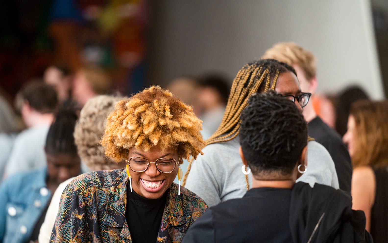 peli2023opn0629kri_004 A woman with curly hair and glasses smiles while talking with others in a lively, crowded indoor setting.