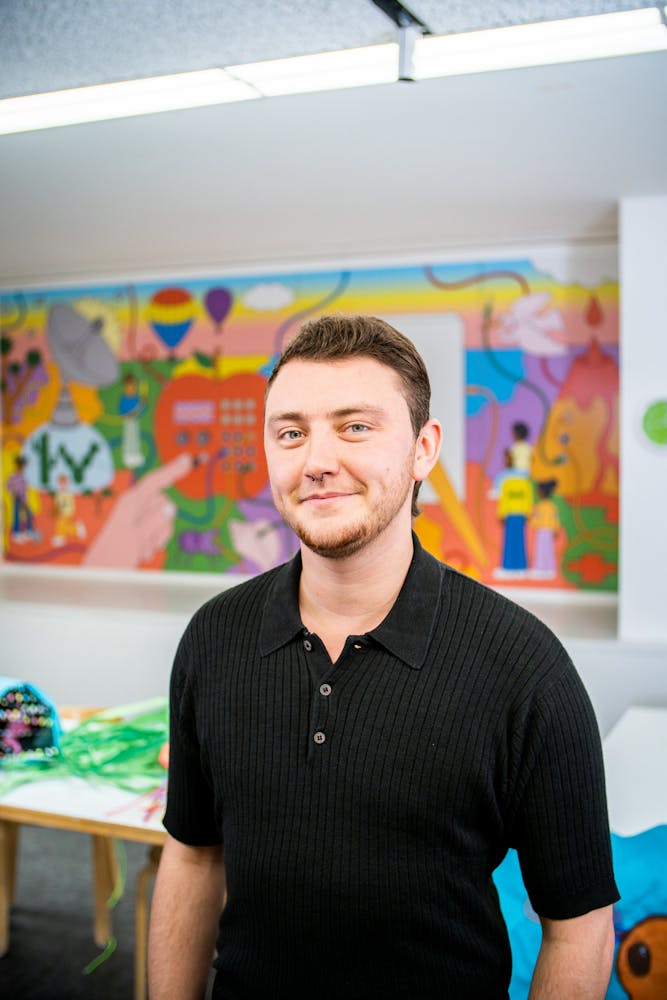 A adult stands in an art classroom and smiles while looking at the viewer.