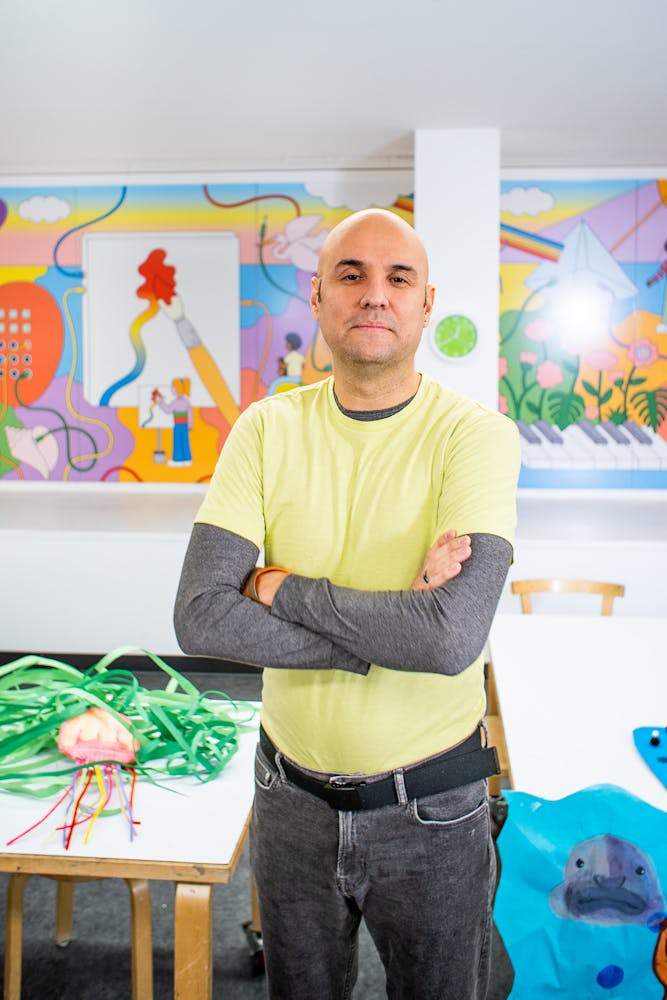 A adult stands in an art classroom and smiles while looking at the viewer.