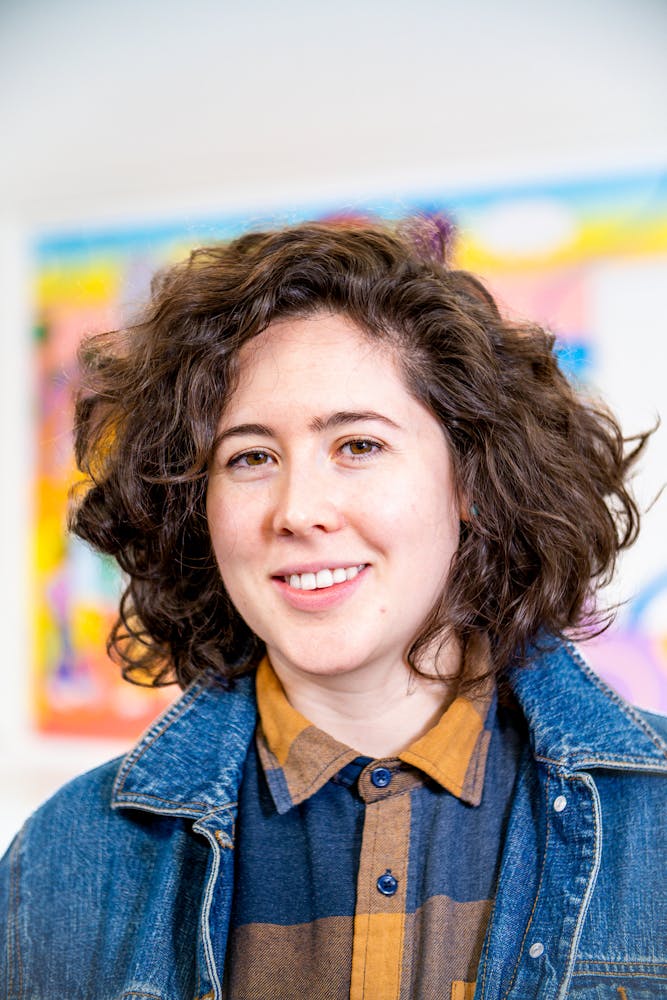 A adult stands in an art classroom and smiles while looking at the viewer.