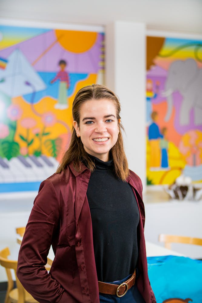 A adult stands in an art classroom and smiles while looking at the viewer.