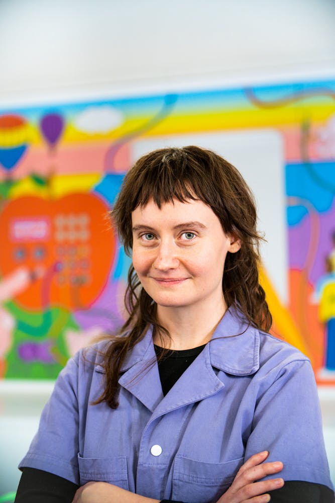 A adult stands in an art classroom and smiles while looking at the viewer.