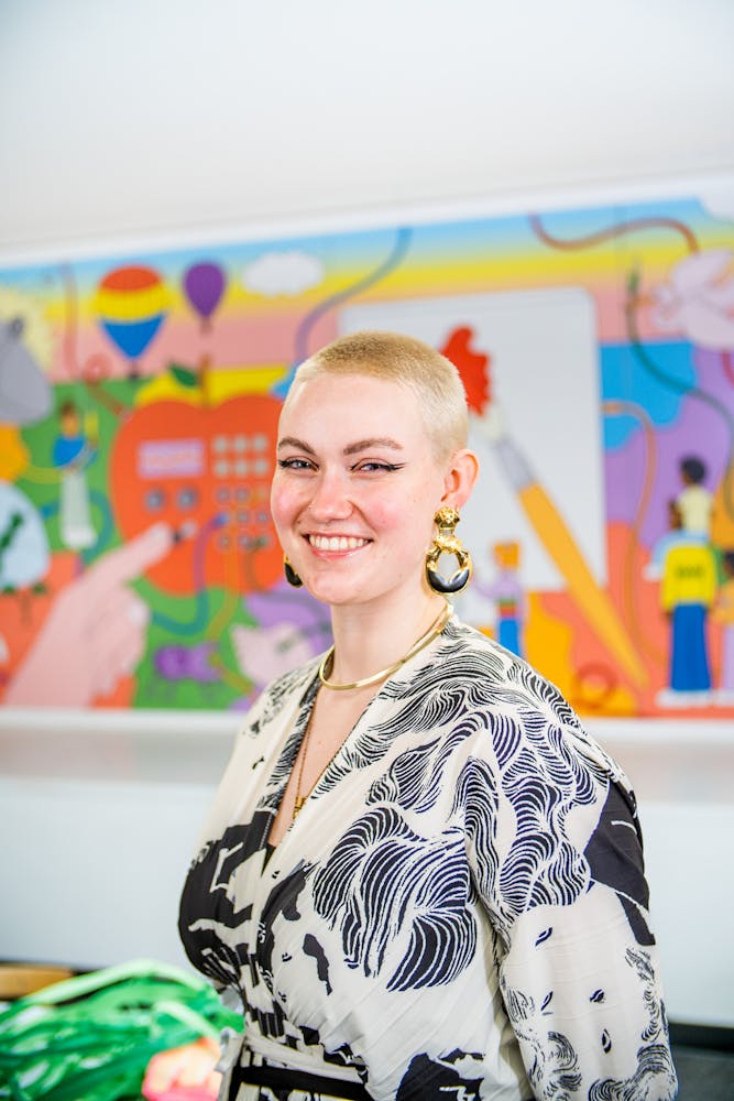 A adult stands in an art classroom and smiles while looking at the viewer.
