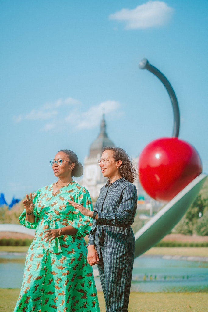 Two women stand and gesture in front of a giant cherry sculpture in a park on a sunny day.