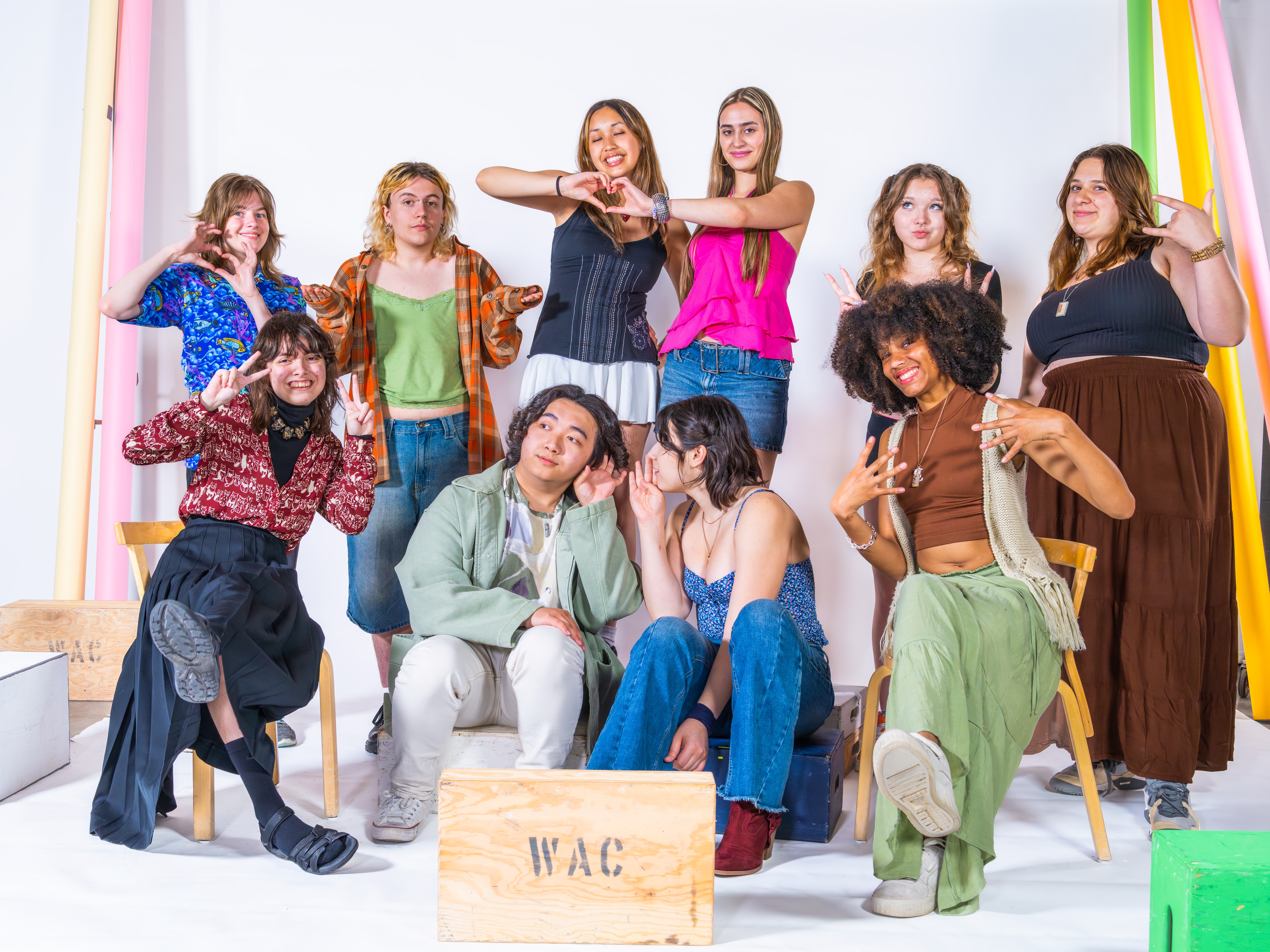 A group of ten teens sit and stand in a photo studio surrounded by colorful paper rolls and a wooden box marked 