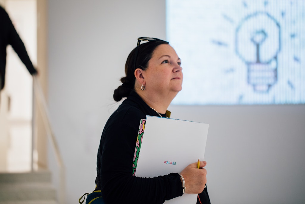 Woman holding folder and pencil, looking up, with a light bulb image on the wall behind her.