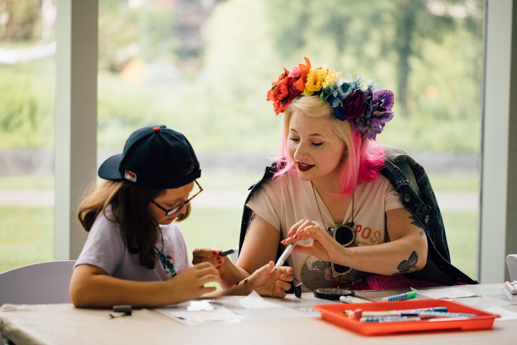 An adult with blonde and pink hair and a flower crown is sitting at a table working on art with a child wearing a backwards baseball cap