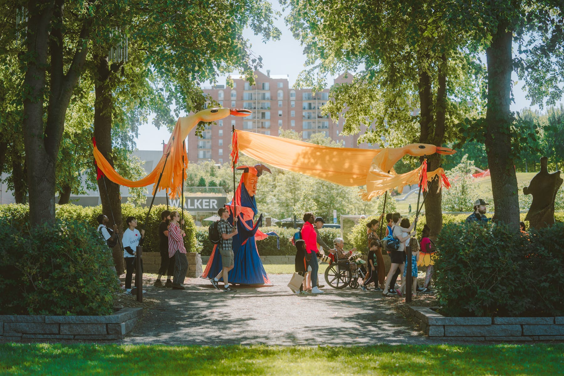 A group of adults and children walk down a sculpture garden path on a sunny summer day while holding large animal creature puppets.
