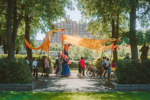 A group of adults and children walk down a sculpture garden path on a sunny summer day while holding large animal creature puppets.