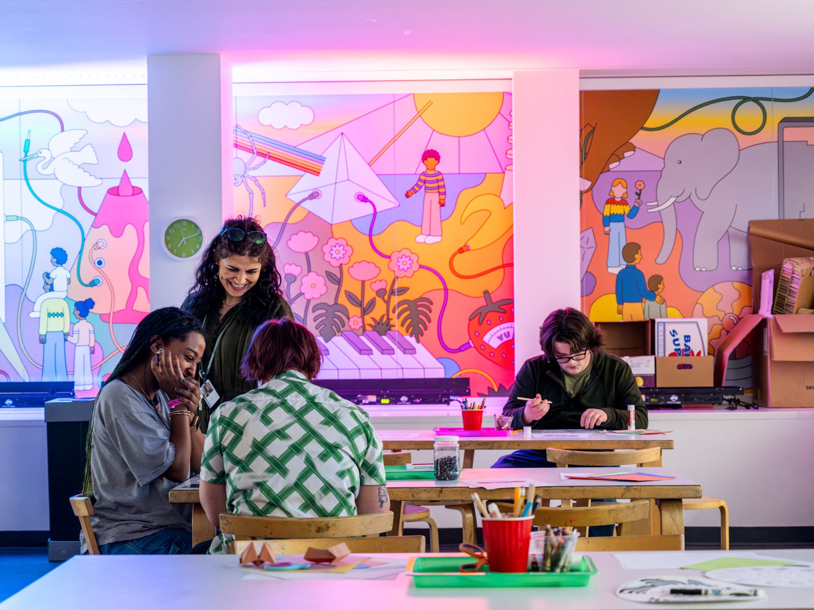 Three adults sit at a tabler in a colorful space, making art.