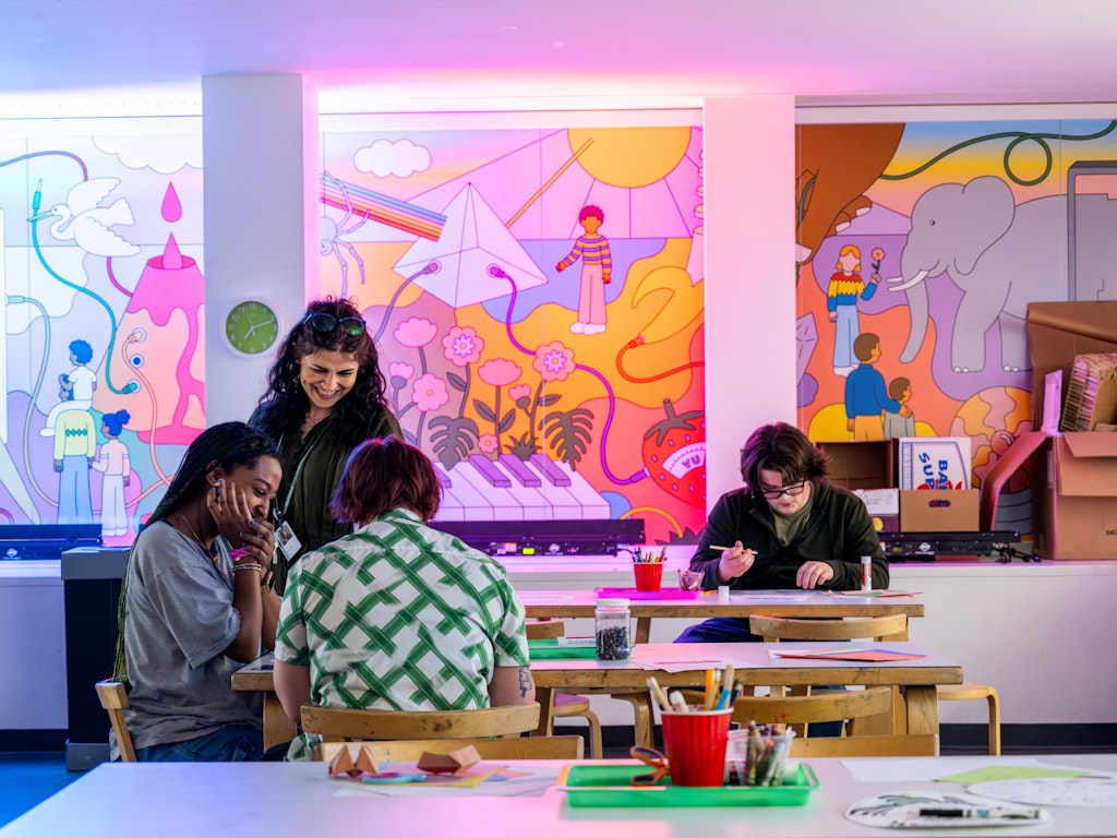 Four people working on art projects at a table in a colorful, brightly decorated classroom with a mural.