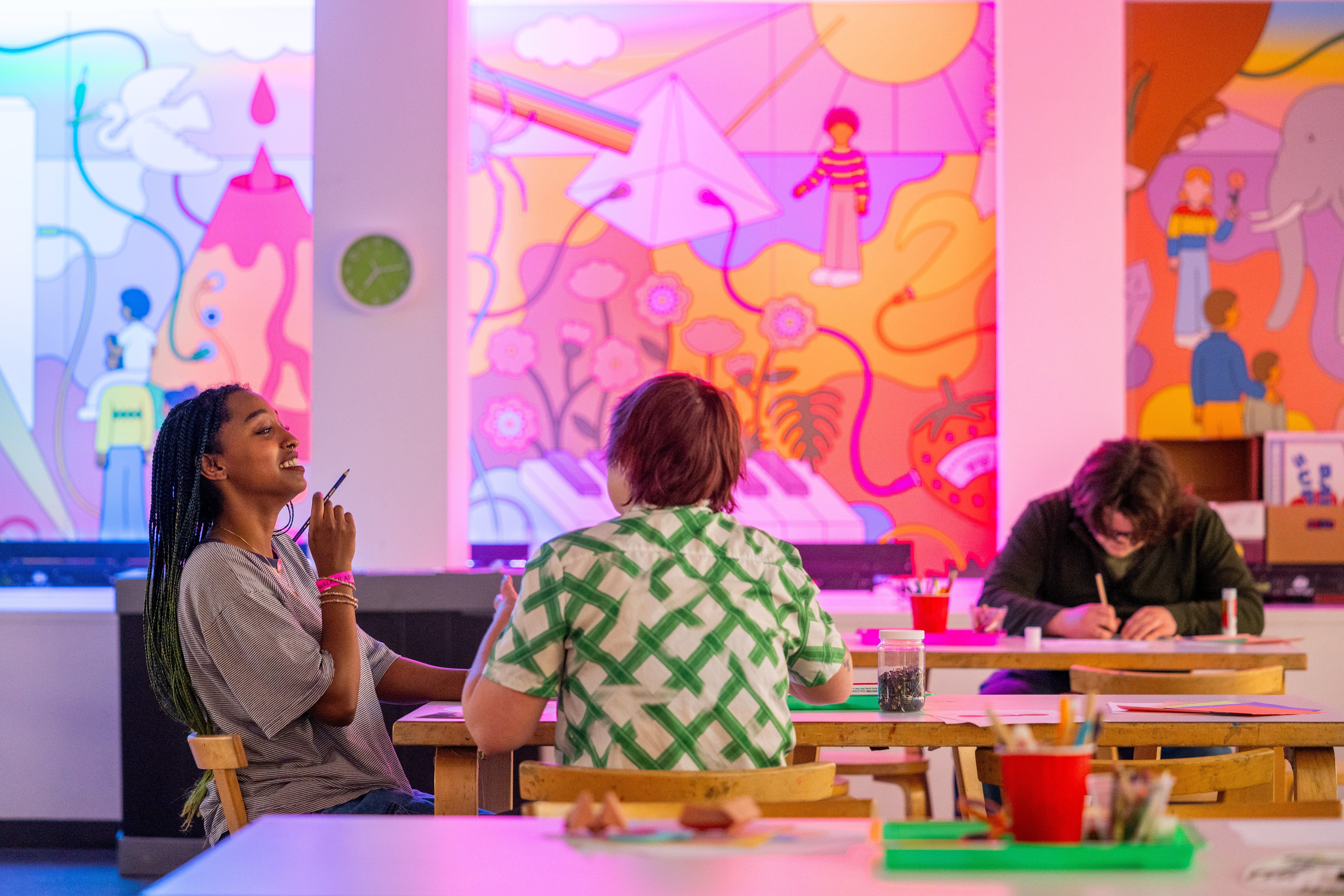 Three adults sit at a tabler in a colorful space, making art.