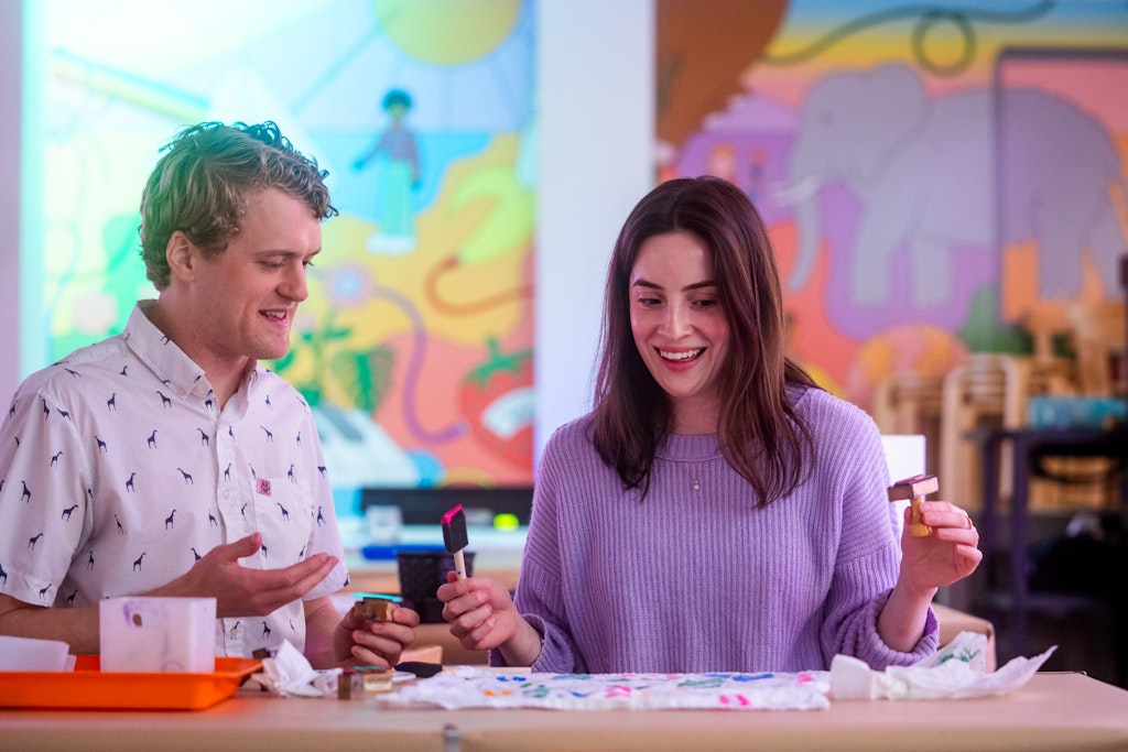 Two adults with light skin and colorful shirts are smiling while they look down at some artwork they are painting.