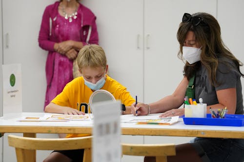 A child with a yellow shirt and a face mask works at a desk with a light skinned adult with long brown hair and a facemask.