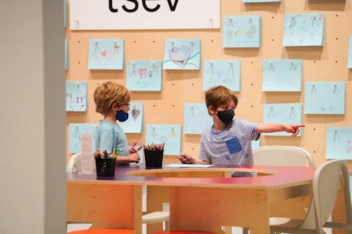 Two children sit at a desk and work on art, one is pointing of camera