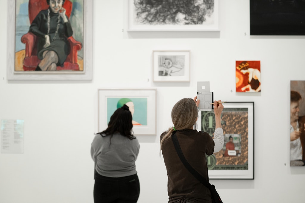 Two people view and photograph artwork on display in a brightly lit art gallery.