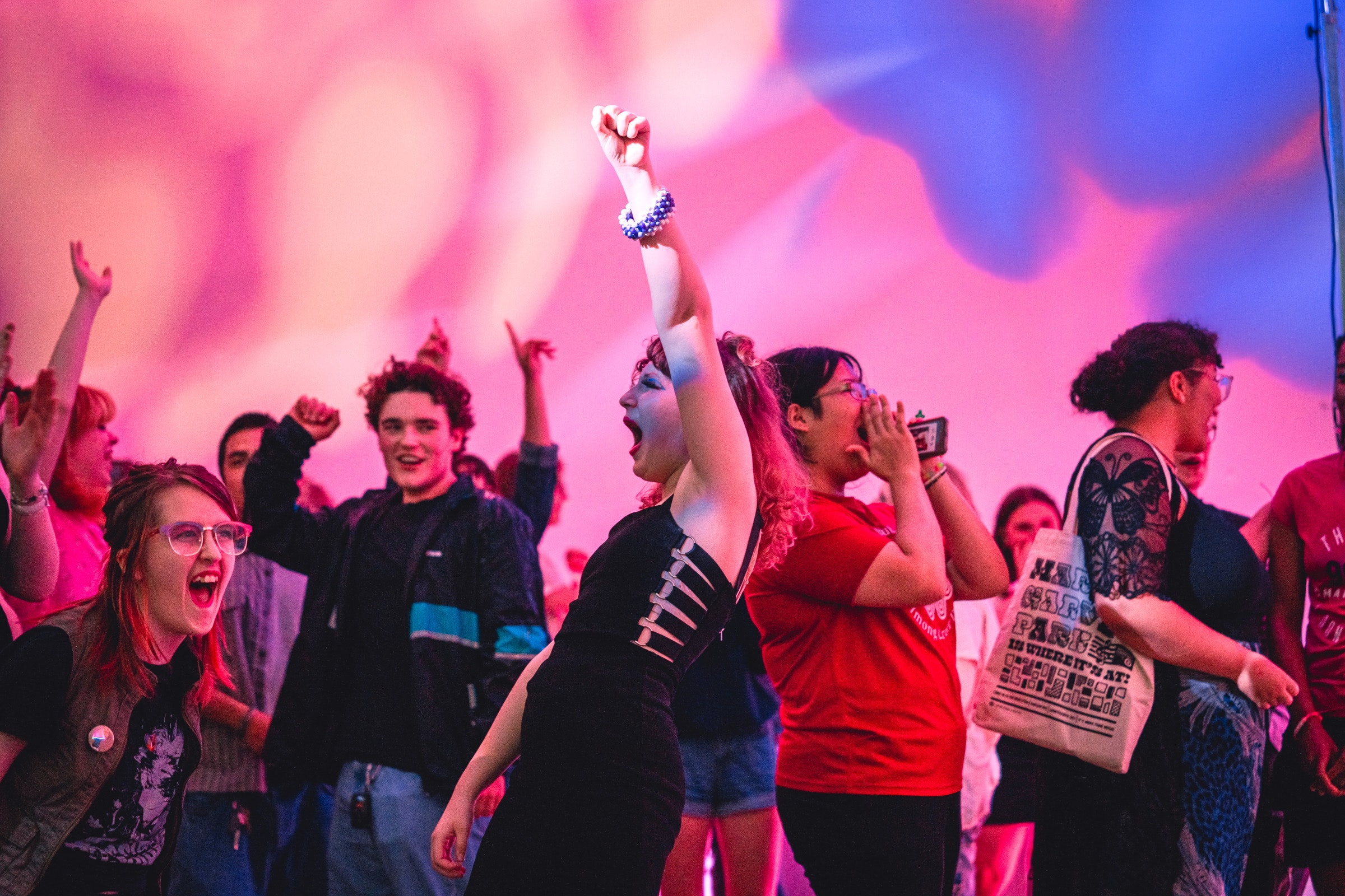 People cheer and celebrate in a colorful, lively crowd, with one person raising their fist in excitement.