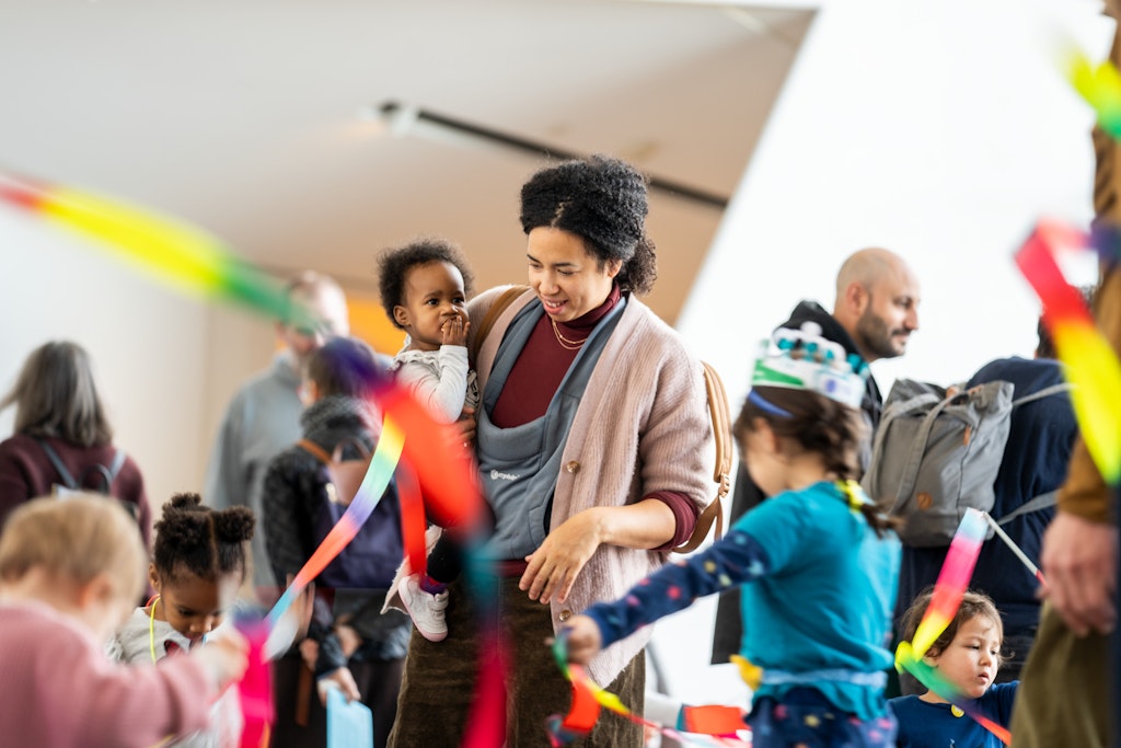 A woman holds a baby, surrounded by children playing with colorful ribbons indoors.