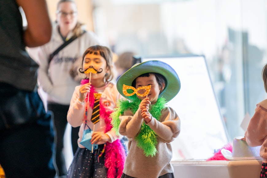 Two children smile while holding up cardboard glasses and mustaches to their faces.
