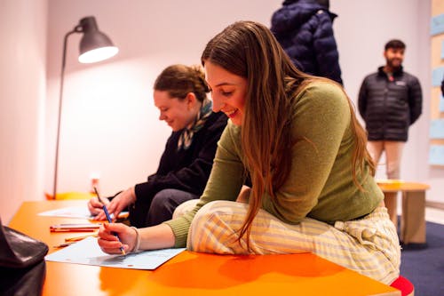 An adult seated at a desk smiles while while drawing.