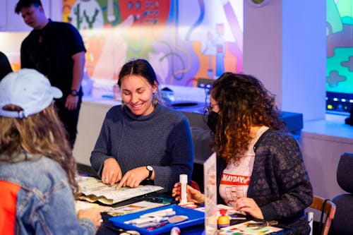 An adult smile to anotherone wearing a face mask as they make 2D artwork together at a table.