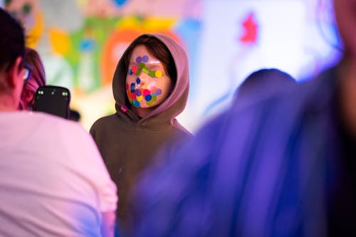 A child with colorful dots all over their face wears a hoodie and posed for a photograpgh.